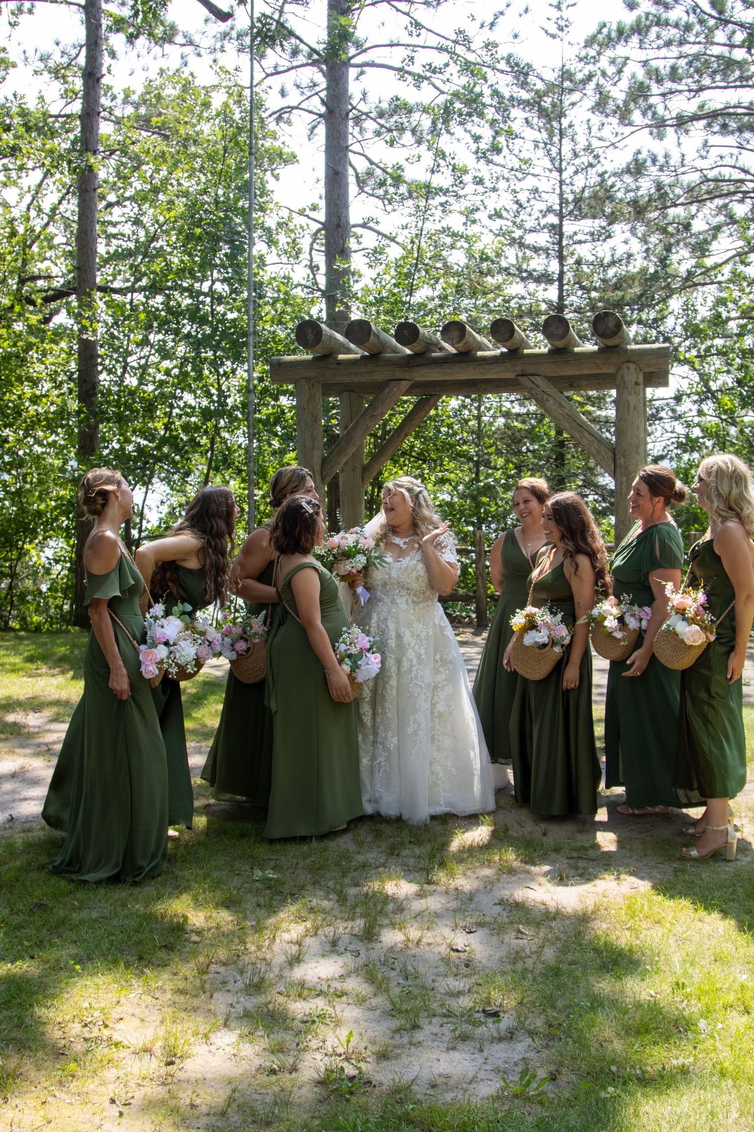 Bridal party in emerald green dresses gathered around bride under wooden pergola in forest setting.