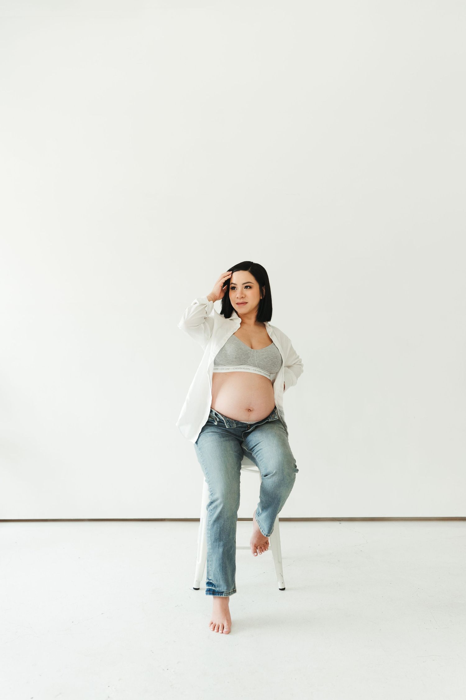 Woman in casual attire poses against white wall in minimalist studio setting.