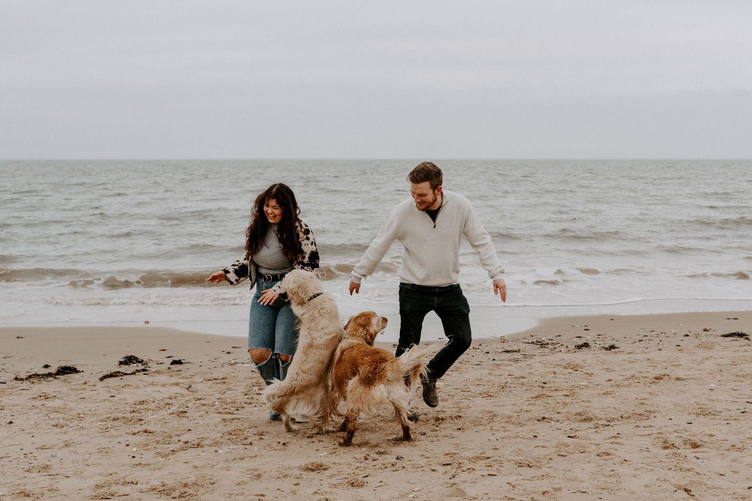A couple run around on the beach with their two golden retriever dogs. They are looking down at their dogs and laughing.