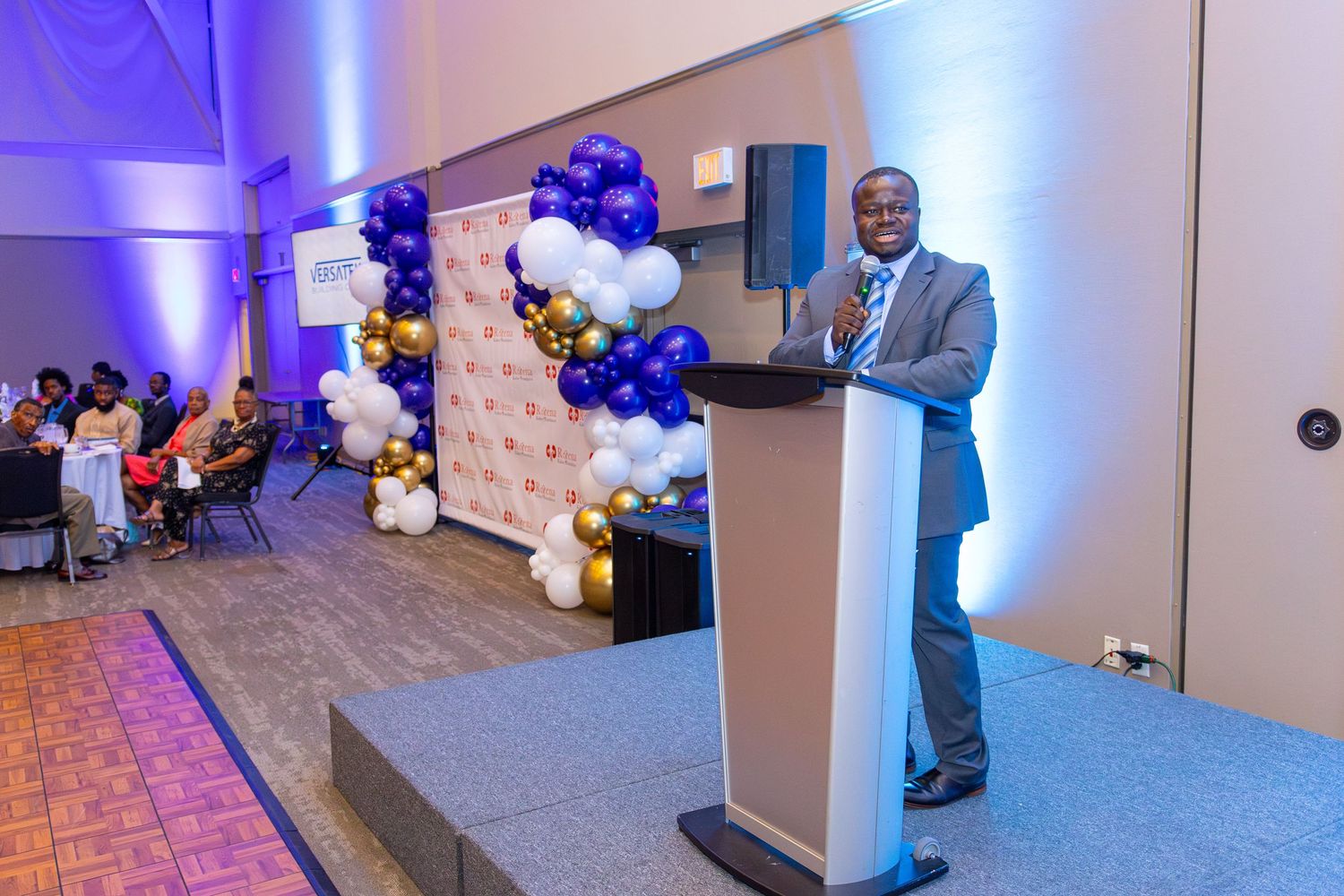 A speaker stands at a podium in an event space decorated with blue and white balloon arrangements.