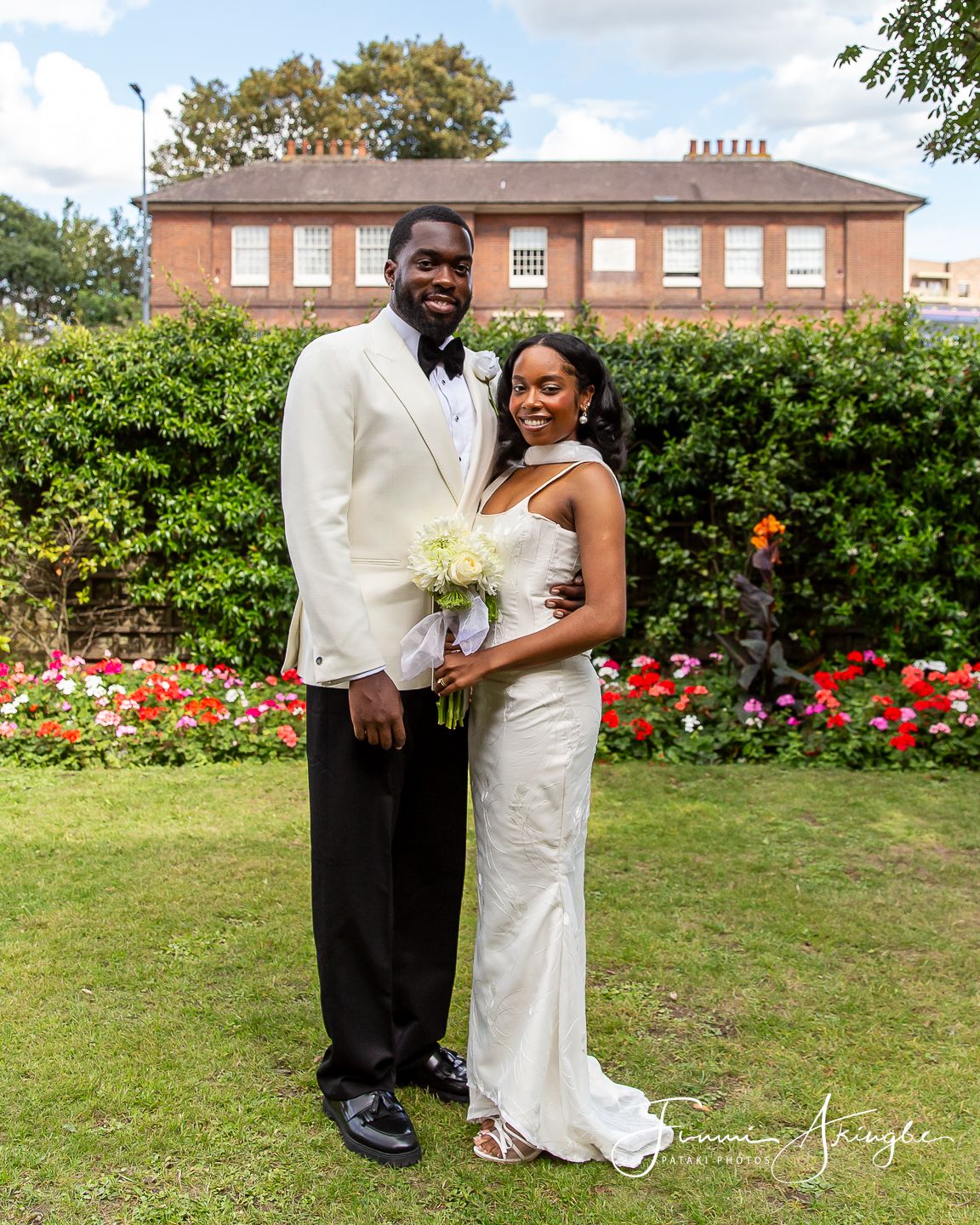 A newly wed bride and groom together in a garden with red roses and green hedges in the background.