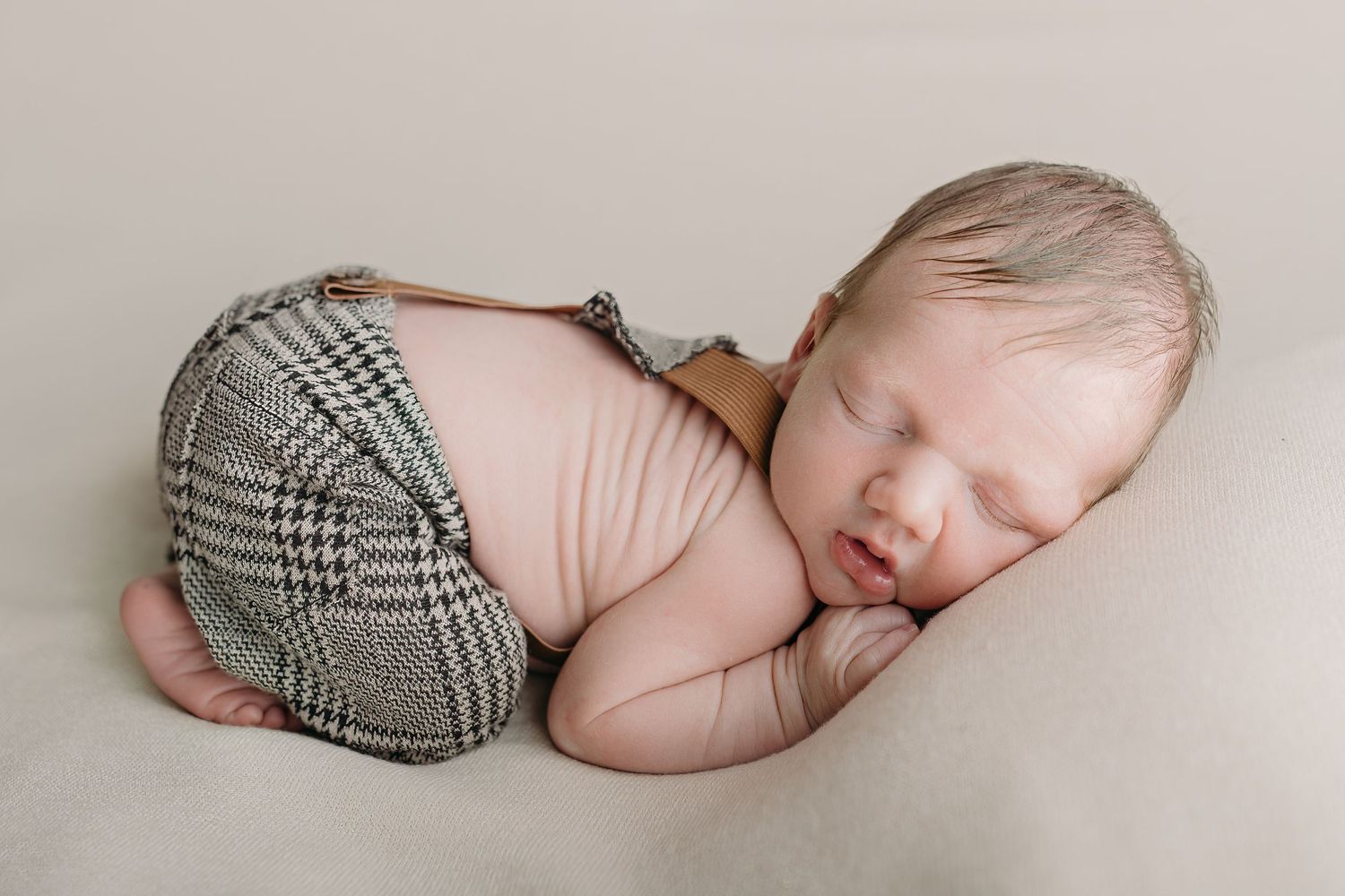 A sleeping newborn baby boy in plaid overalls lies peacefully on a white backdrop during newborn photography session in Tampa