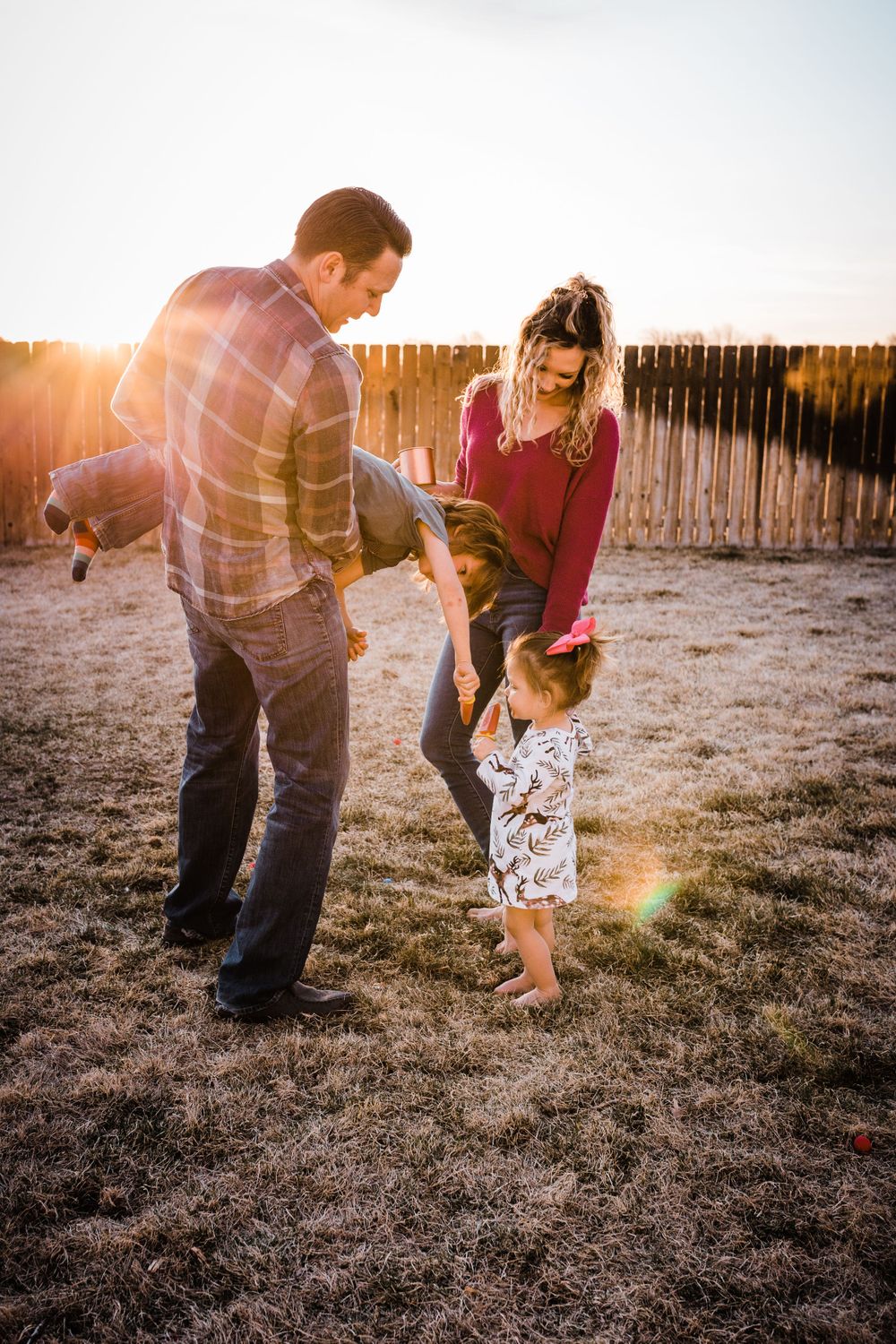 Family playing together in backyard at sunset with warm golden light and wooden fence in background.