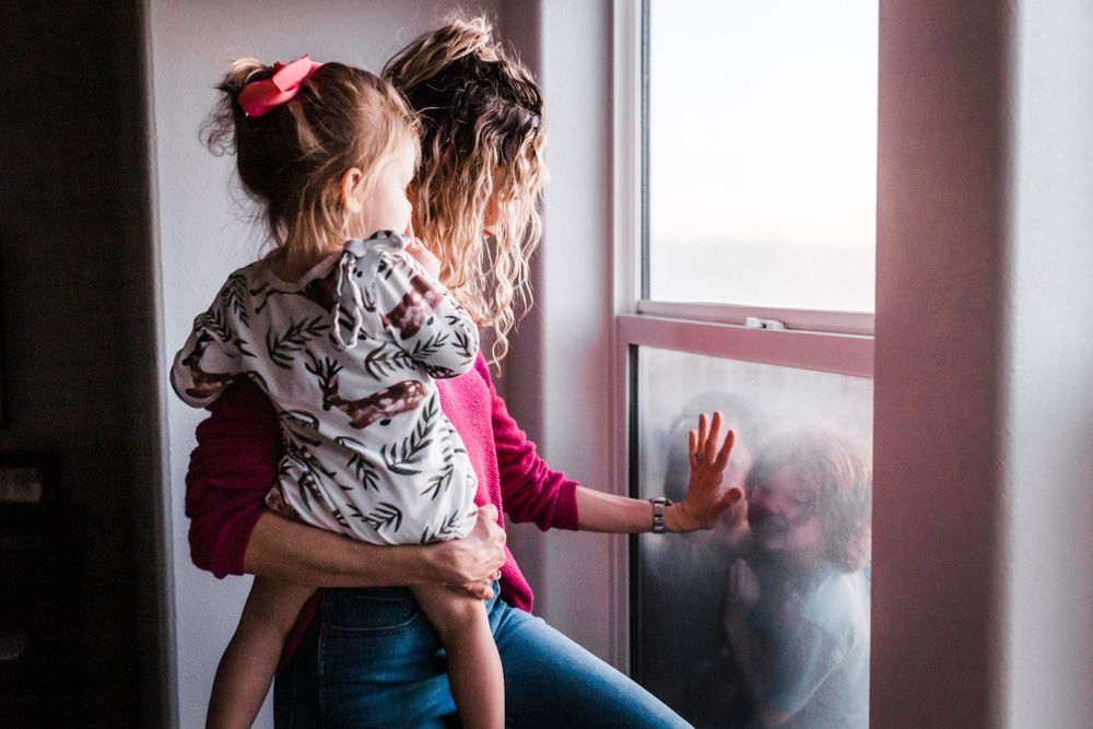 A caregiver holds a child at a window during a quiet moment on a dreary day.