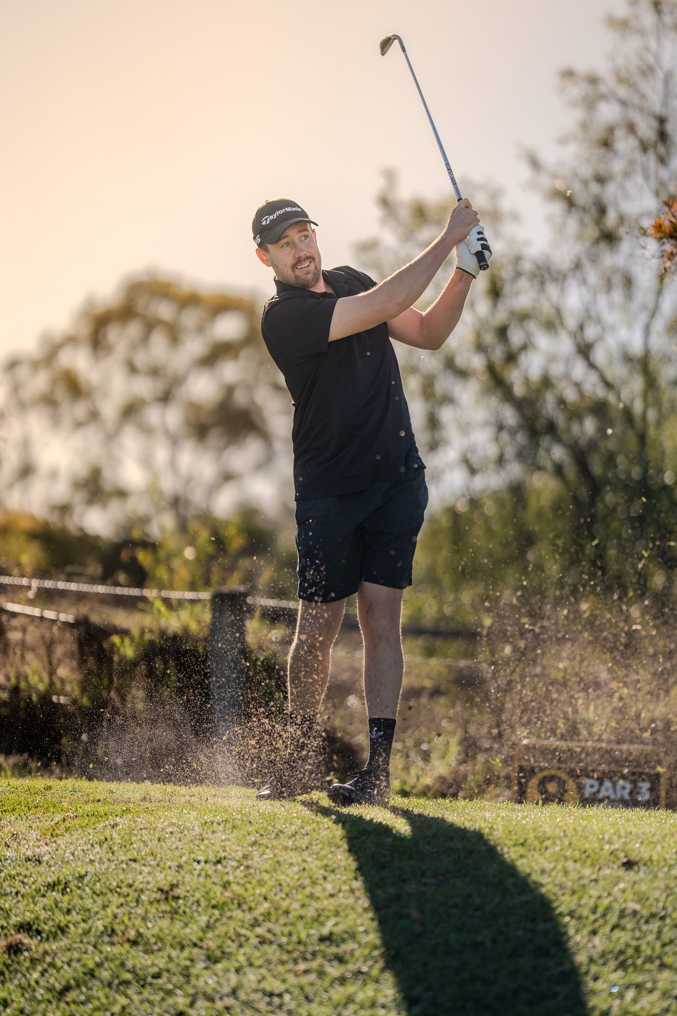 Golfer wearing black apparel swings club on course during sunset as dirt sprays from impact at tee box.