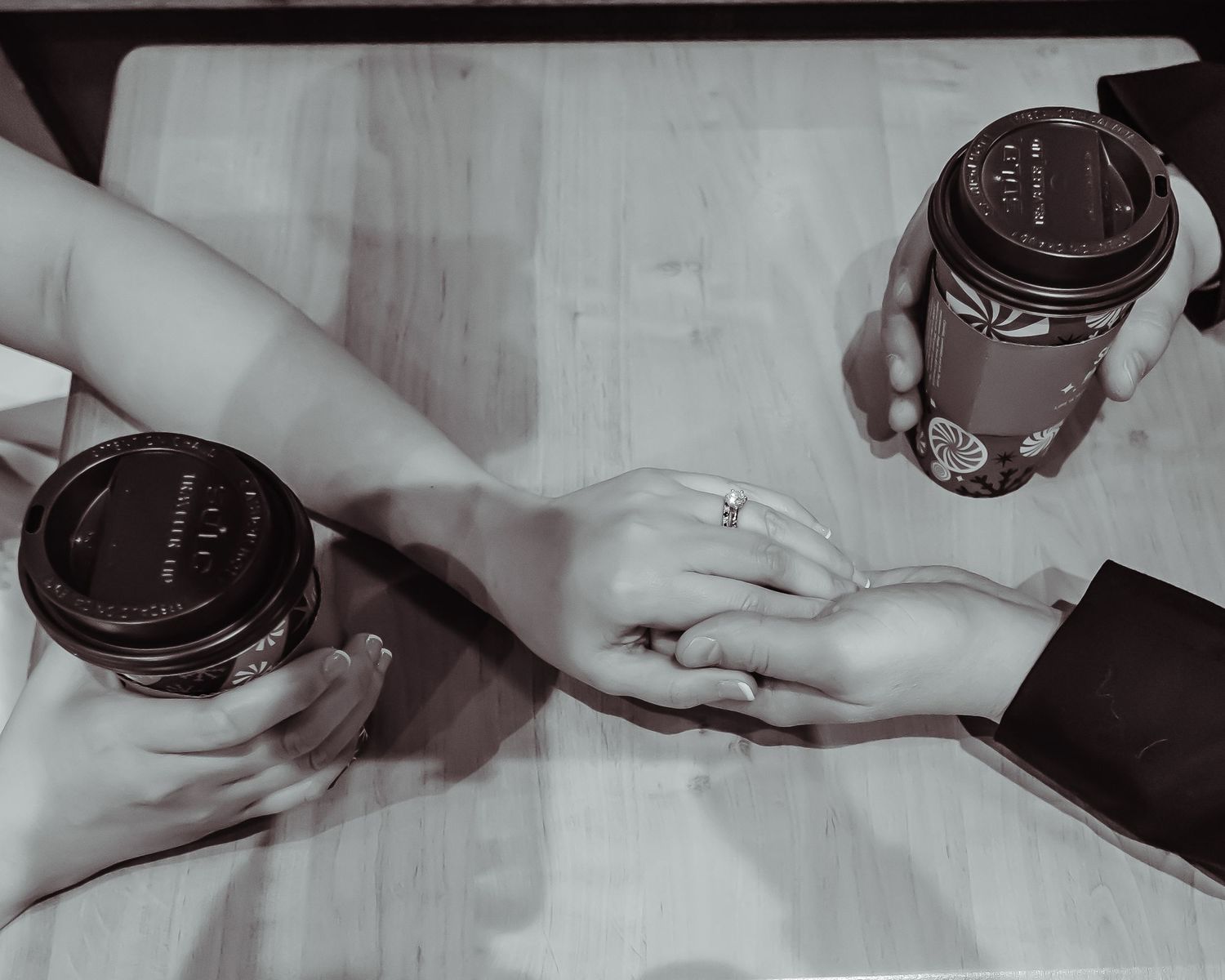 Black and white photo of two hands holding coffee cups on a table, with fingers touching intimately.