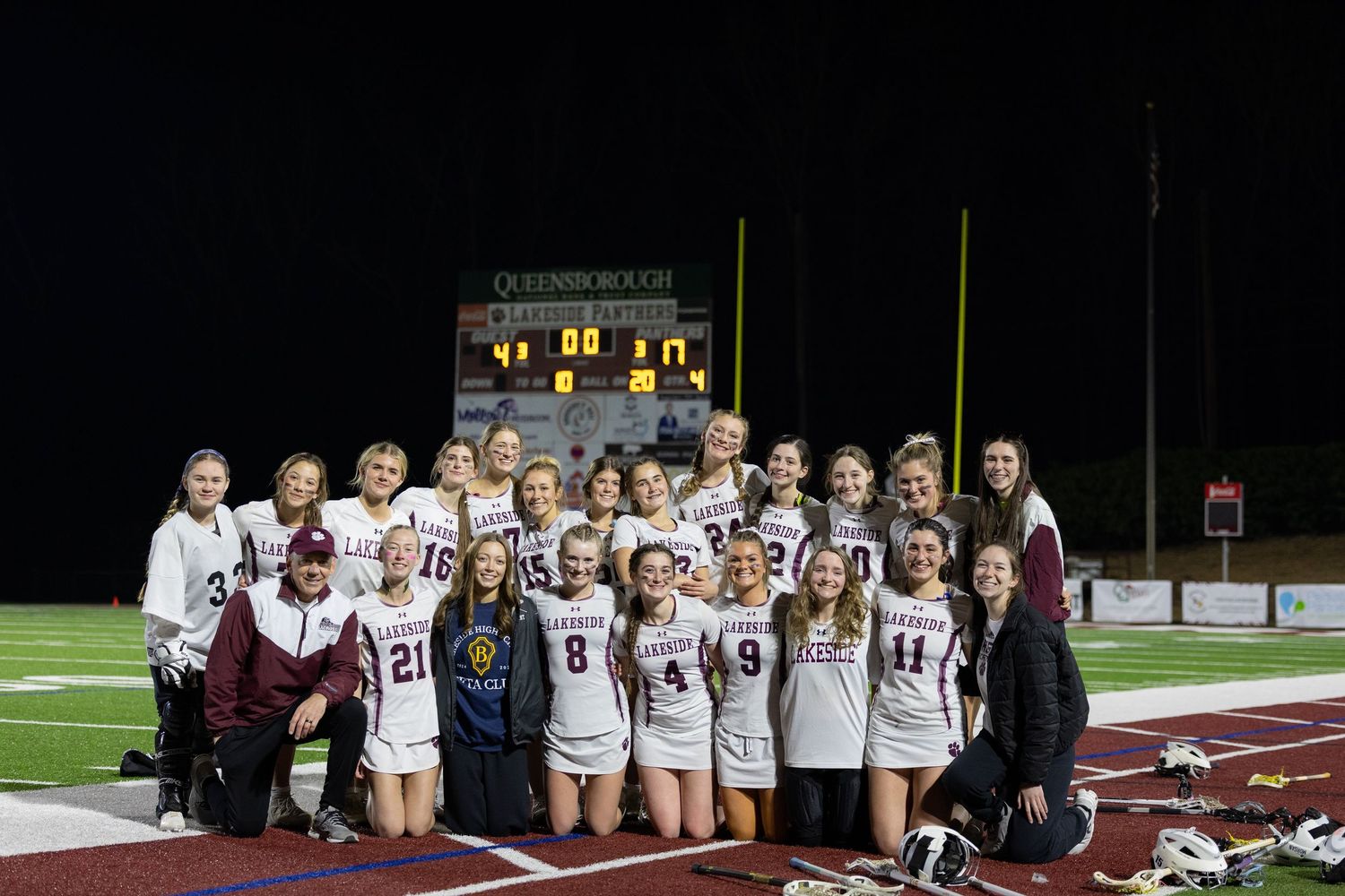 Large group poses beneath electronic scoreboard with sponsor logos at night.