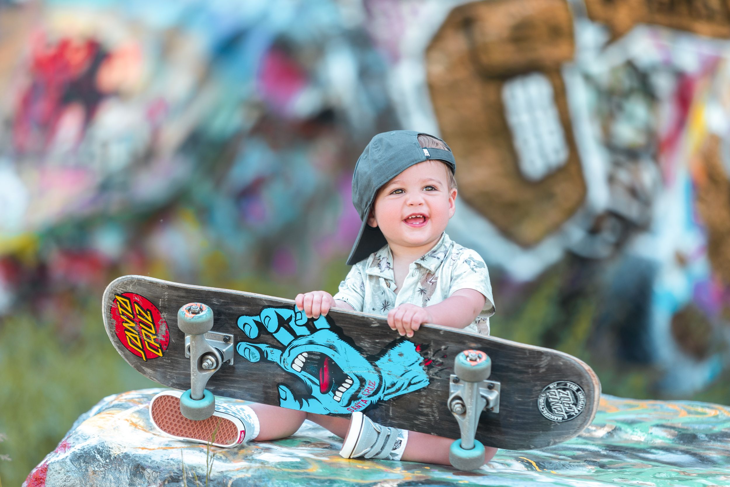 Small child in blue beanie sits with skateboard against colorful graffiti background outdoors.