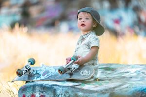 Child in backwards cap looking up while sitting with skateboard on outdoor surface at golden hour.