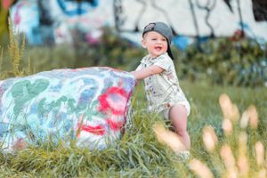 Small child in white romper plays outdoors near colorful painted rock with grass and foliage in background.