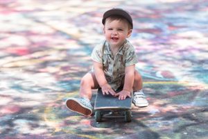 Child in casual wear sitting on skateboard against colorful tie-dye backdrop.