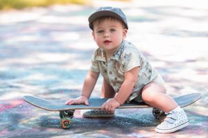 A toddler in a baseball cap crouches on a skateboard while wearing patterned shorts and sneakers.