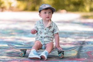 Young child in blue cap and patterned onesie sitting on a skateboard wearing blue sneakers.