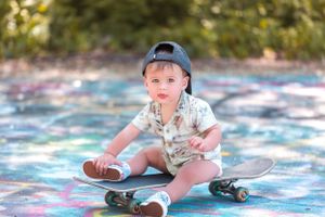 Small child in baseball cap sitting on a decorated skateboard against a colorful painted surface on a sunny day.