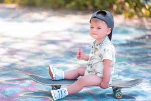 A small child sits on a skateboard wearing a baseball cap and white t-shirt against a blurred outdoor background.
