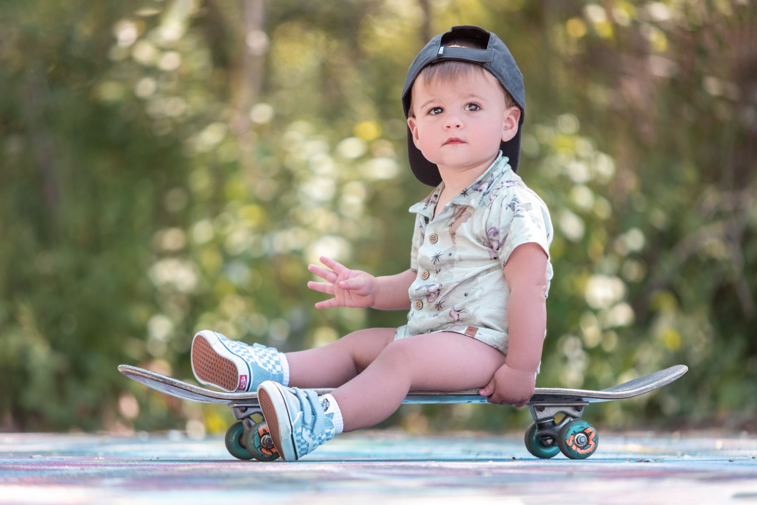 Young child wearing a baseball cap and floral shirt sits on a skateboard in a sunny outdoor setting.