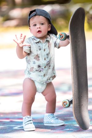 Small child in baseball cap and patterned onesie standing next to a skateboard outdoors.