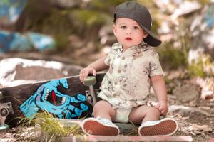 Small child in a baseball cap sits on dirt ground next to a blue and black backpack with casual summer attire.