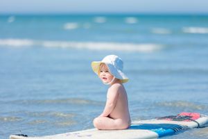 Child in sunhat sits on paddleboard in turquoise ocean waters on bright sunny day.