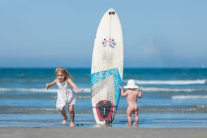 A decorative surfboard with blue details stands upright on a sandy beach by the ocean.