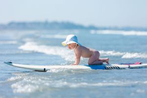 Small baby wearing blue hat learning to surf on a surfboard in gentle ocean waves at the beach.