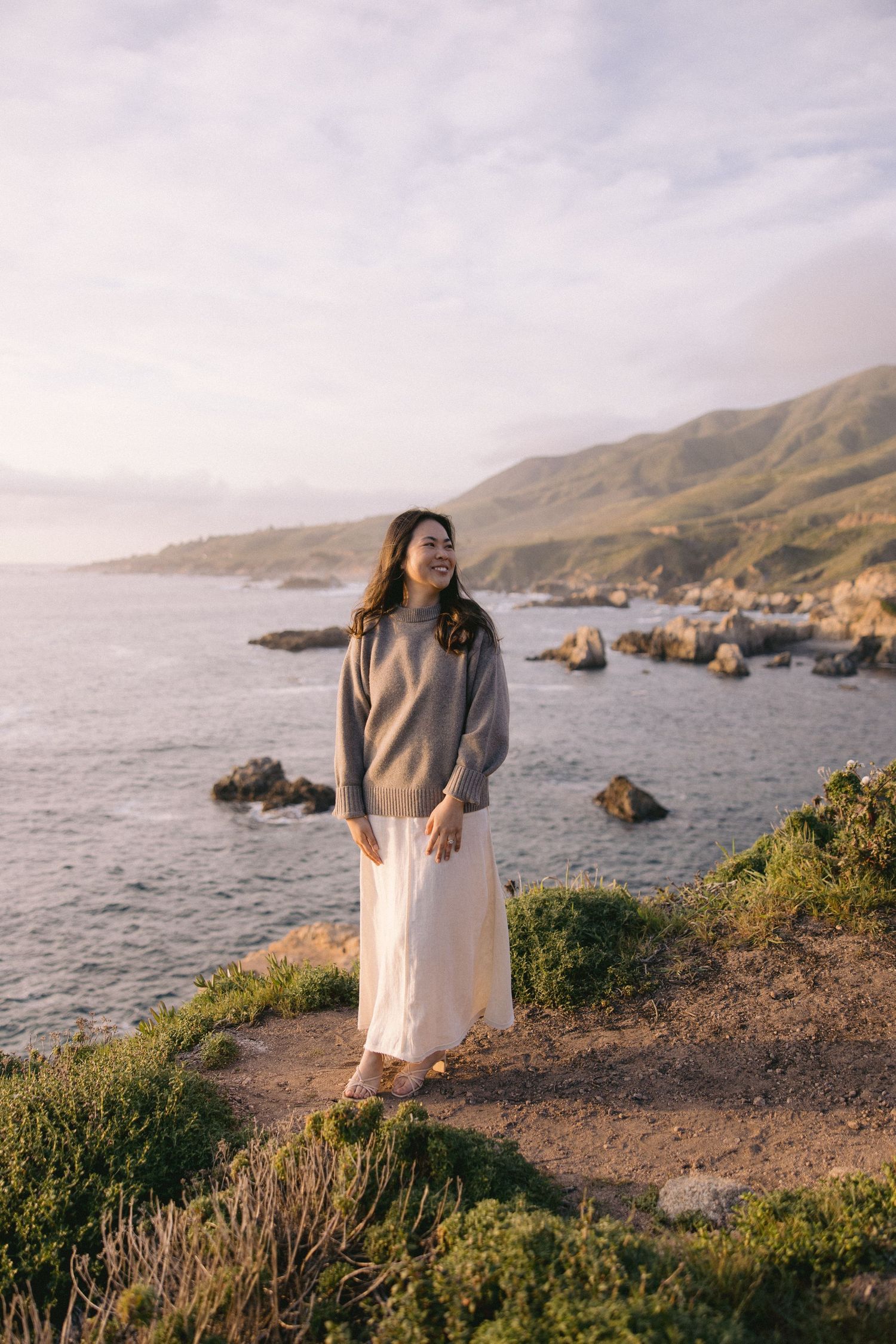 Someone in a cozy sweater and white skirt stands on a coastal cliff overlooking Big Sur's rocky shoreline at sunset.