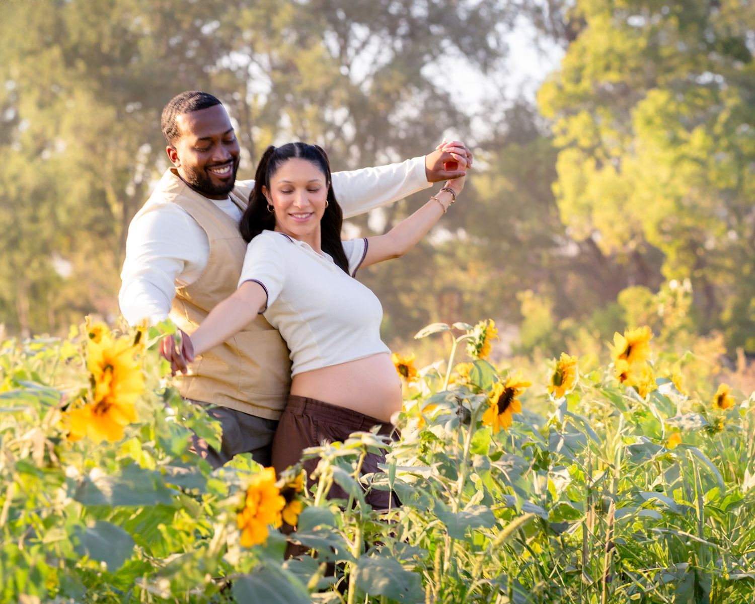 A couple poses together in a sunflower field during maternity photo session, with autumn trees in the background.