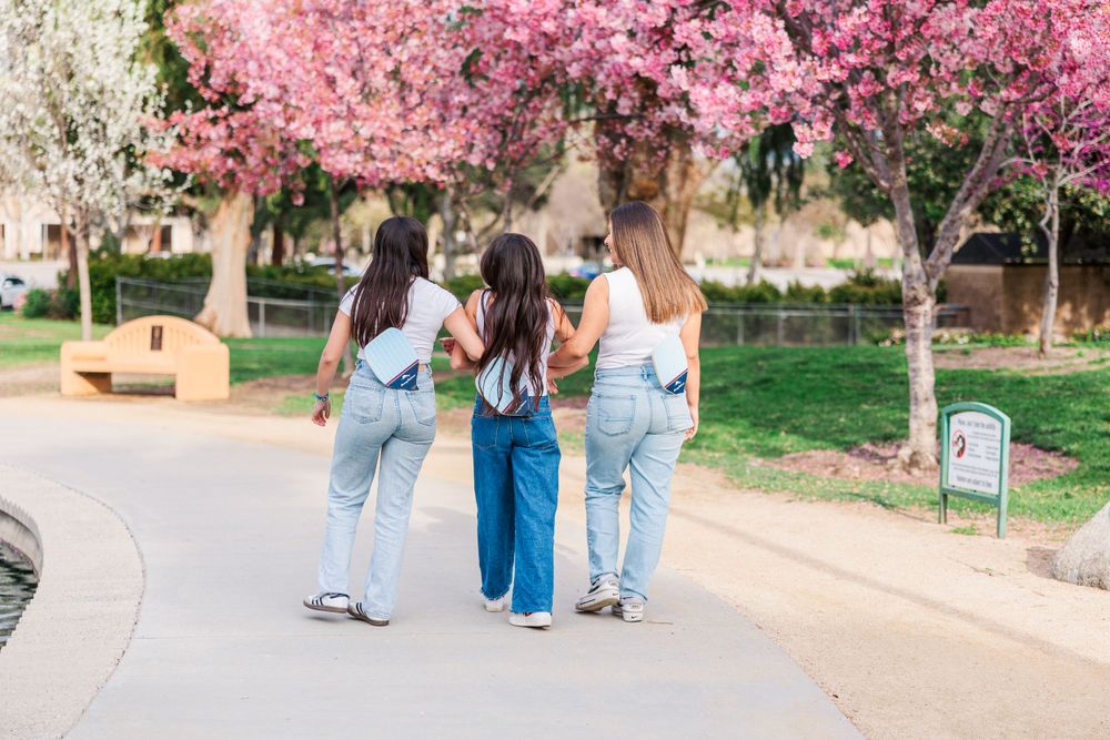 Three friends walk arm in arm down a path beneath blooming pink cherry blossom trees in spring.Simi Valley Photographer