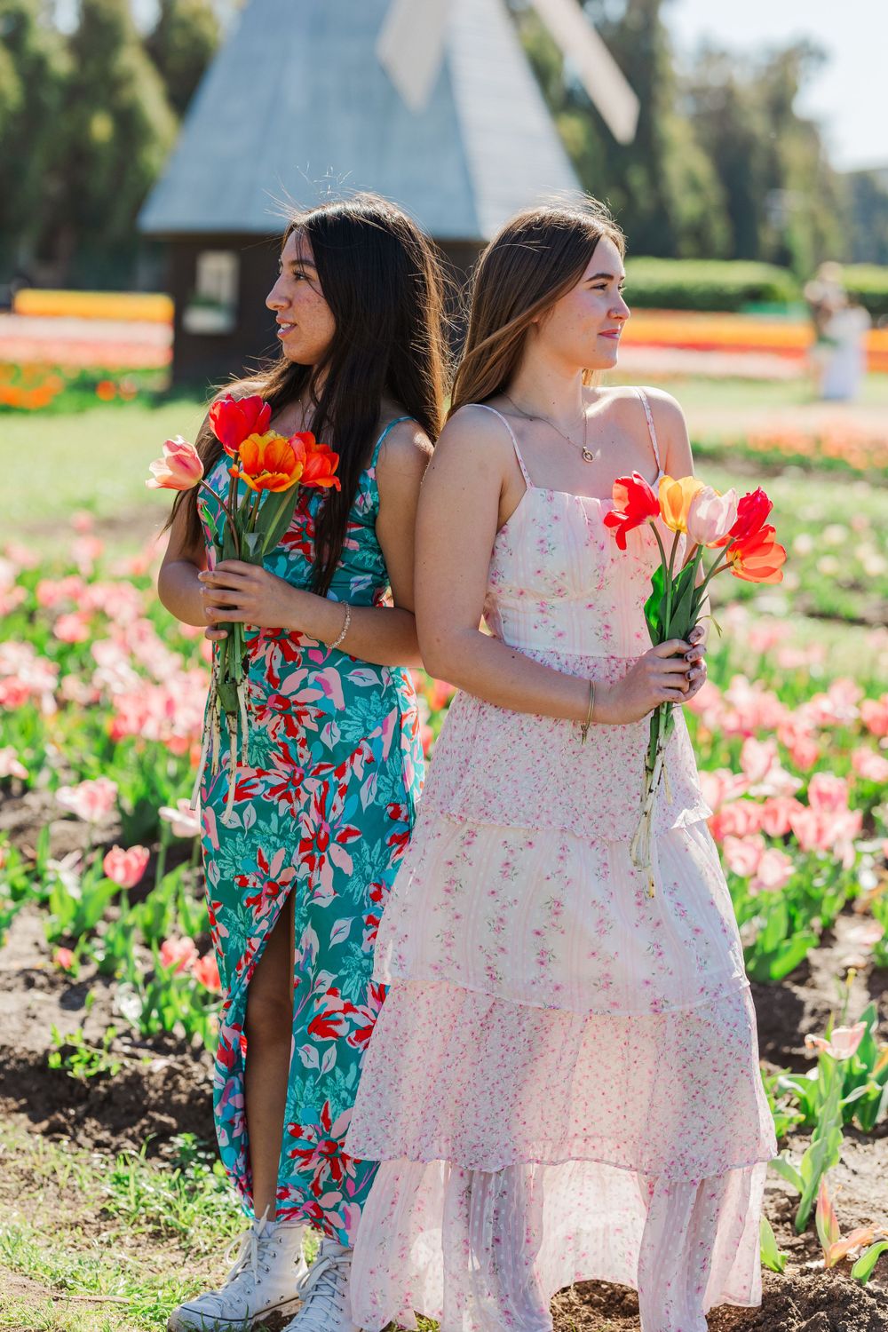 Two women in floral dresses pose back-to-back holding bright tulips in a sunny garden filled with colorful flowers.Simi Valley Senior Photo