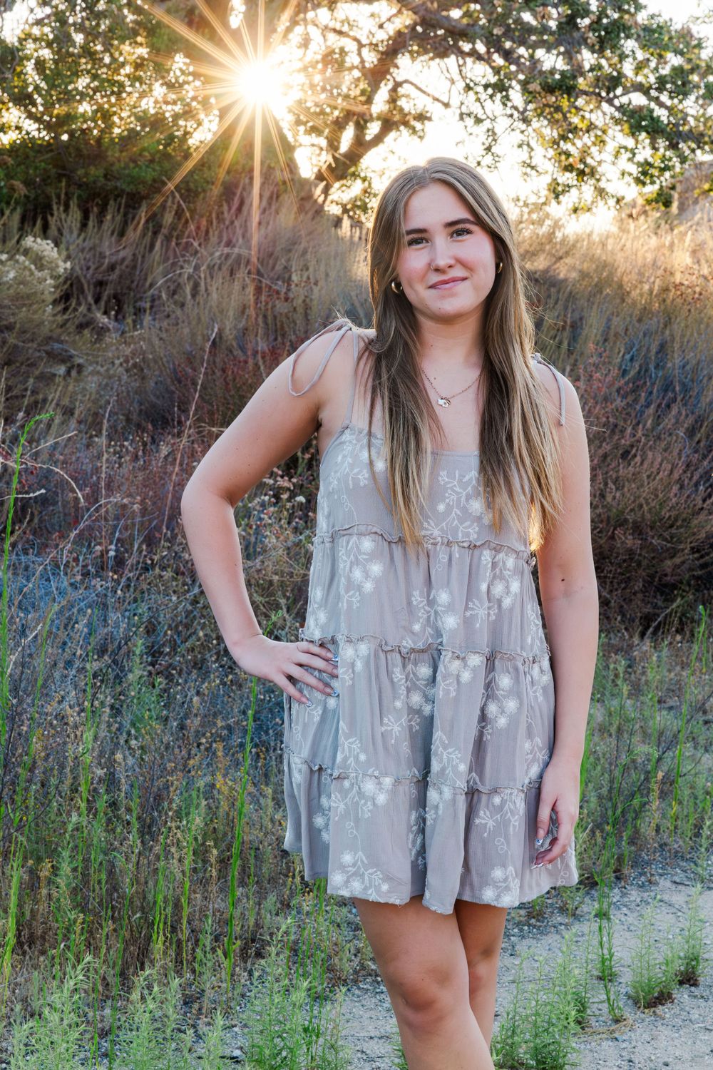 Woman in tiered gray sundress poses in golden sunlit field at sunset against trees.Simi Valley Senior Photographer
