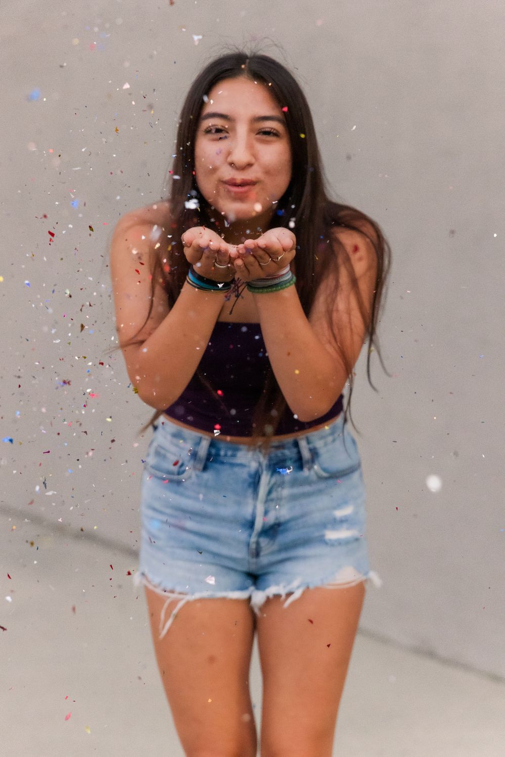 Someone in denim shorts and black top blows glitter and confetti against a gray wall for a fun celebratory photo.Simi Valley Senior Photogra