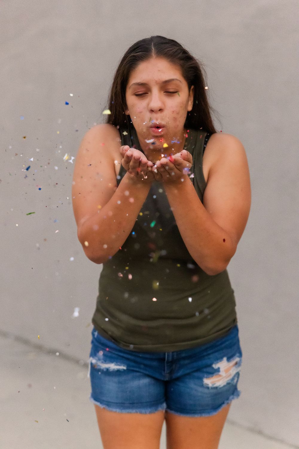 Someone wearing an olive tank top and denim shorts blows sparkly confetti from their hands against a gray background.Simi Valley Photographe