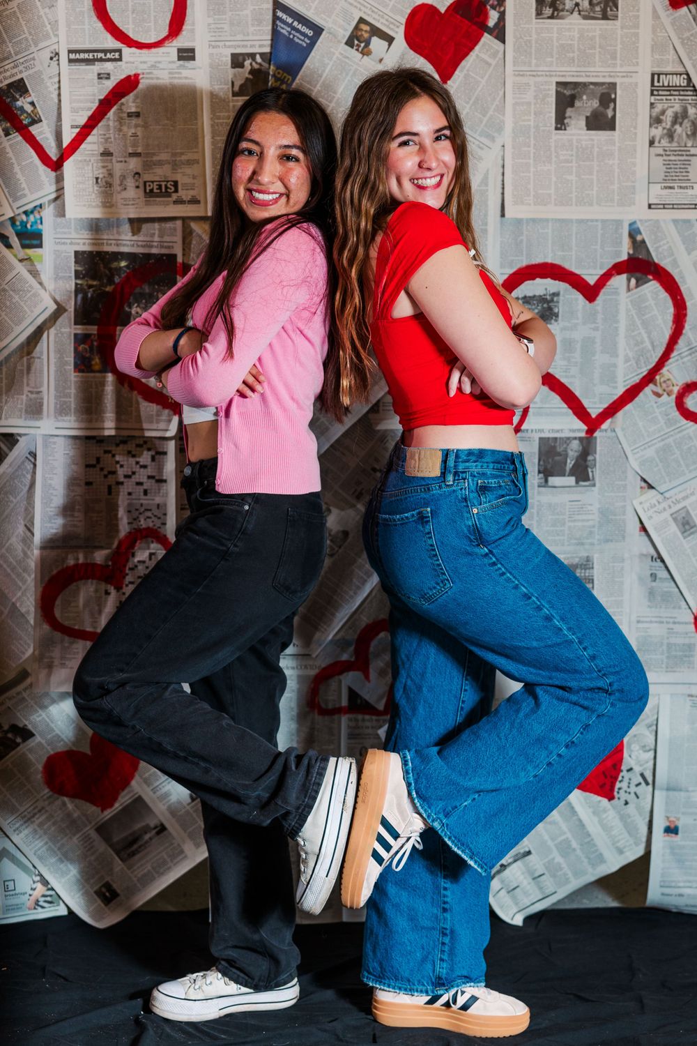 Two friends in pink and red crop tops and jeans pose back-to-back against a newspaper wall decorated with red hearts.Simi Valley Senior Phot