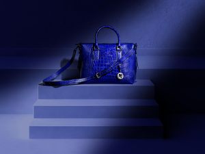 Royal blue crocodile-embossed leather handbag with gold hardware displayed on blue geometric steps.