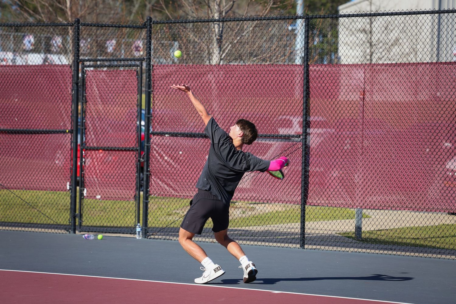 Tennis player serving the ball on an outdoor court with red fence and trees in the background.