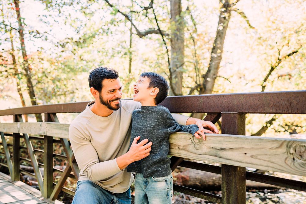 A parent and child share a tender moment together while sitting on a wooden bridge in an autumn forest setting.