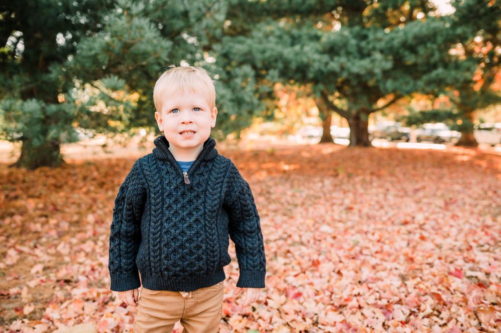 A small figure in a navy cable knit sweater stands amid fallen autumn leaves in a park setting.