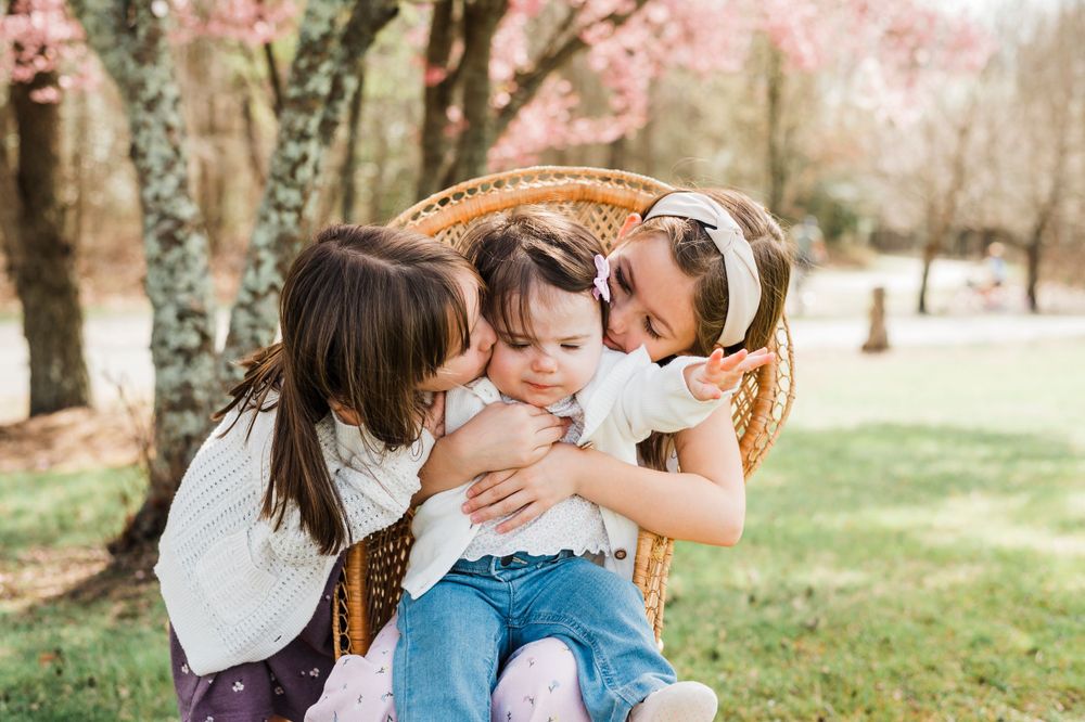 Children laugh together while sitting on a vintage style wicker chair outdoors under pink blossoming trees in spring.