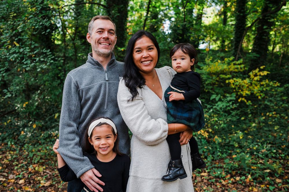 A family of four poses together for an outdoor portrait in a lush green wooded setting during autumn.