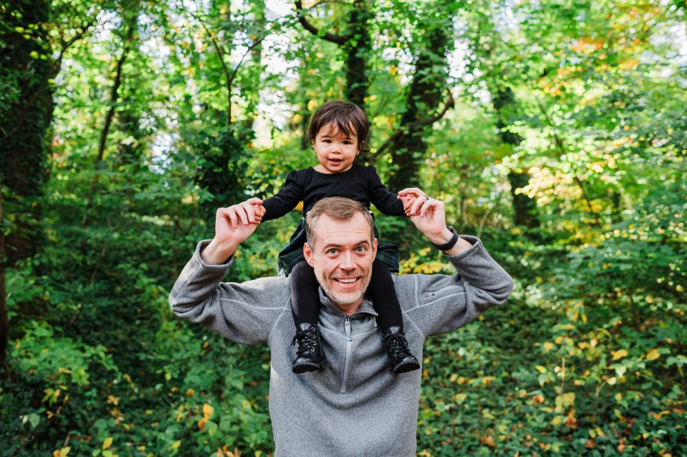 A person wearing a gray hoodie gives a shoulder ride through a lush green forest on a sunny day.