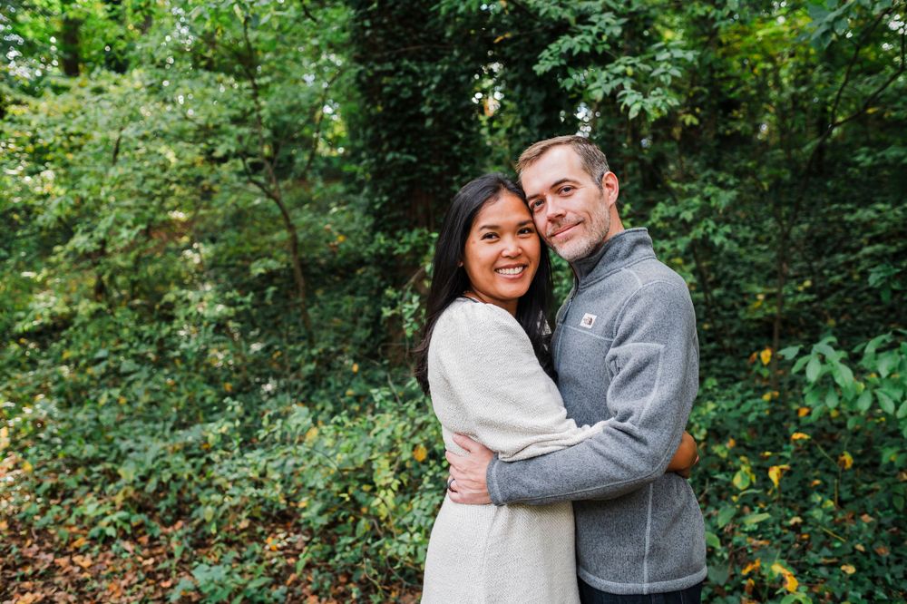 A happy couple embraces outdoors while wearing cozy sweaters against a lush green forest backdrop.