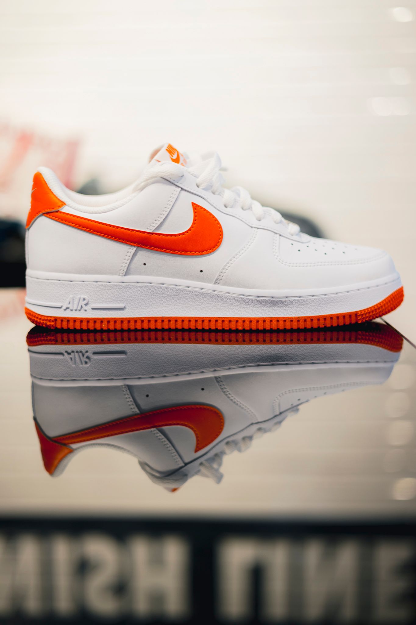 Nike Air Force 1 sneaker in white and orange colorway with mirror reflection on glossy surface.