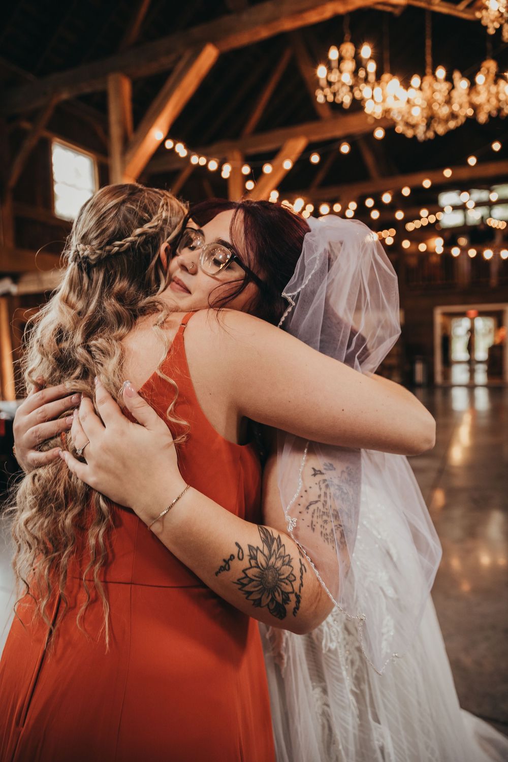 Emotional embrace captured at indoor rustic wedding venue with warm string lights and chandeliers creating romantic ambiance.