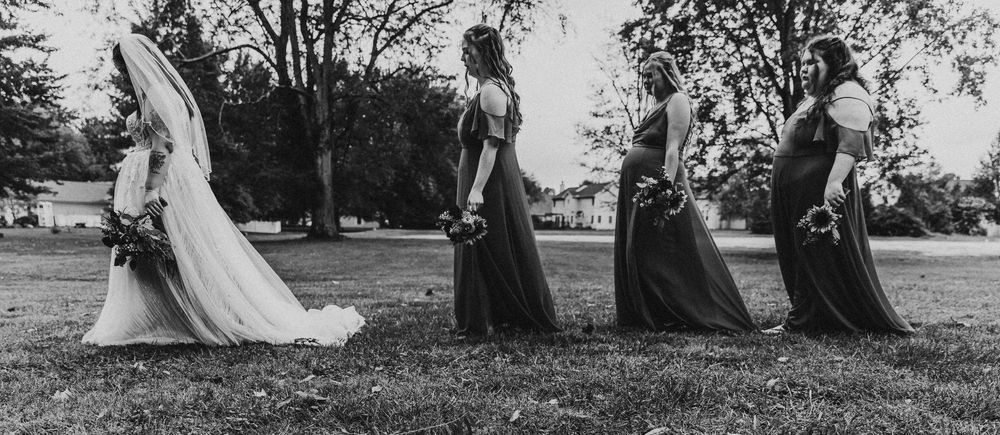A bridal party walks in line across a grassy field in black and white, with a trailing wedding dress visible.