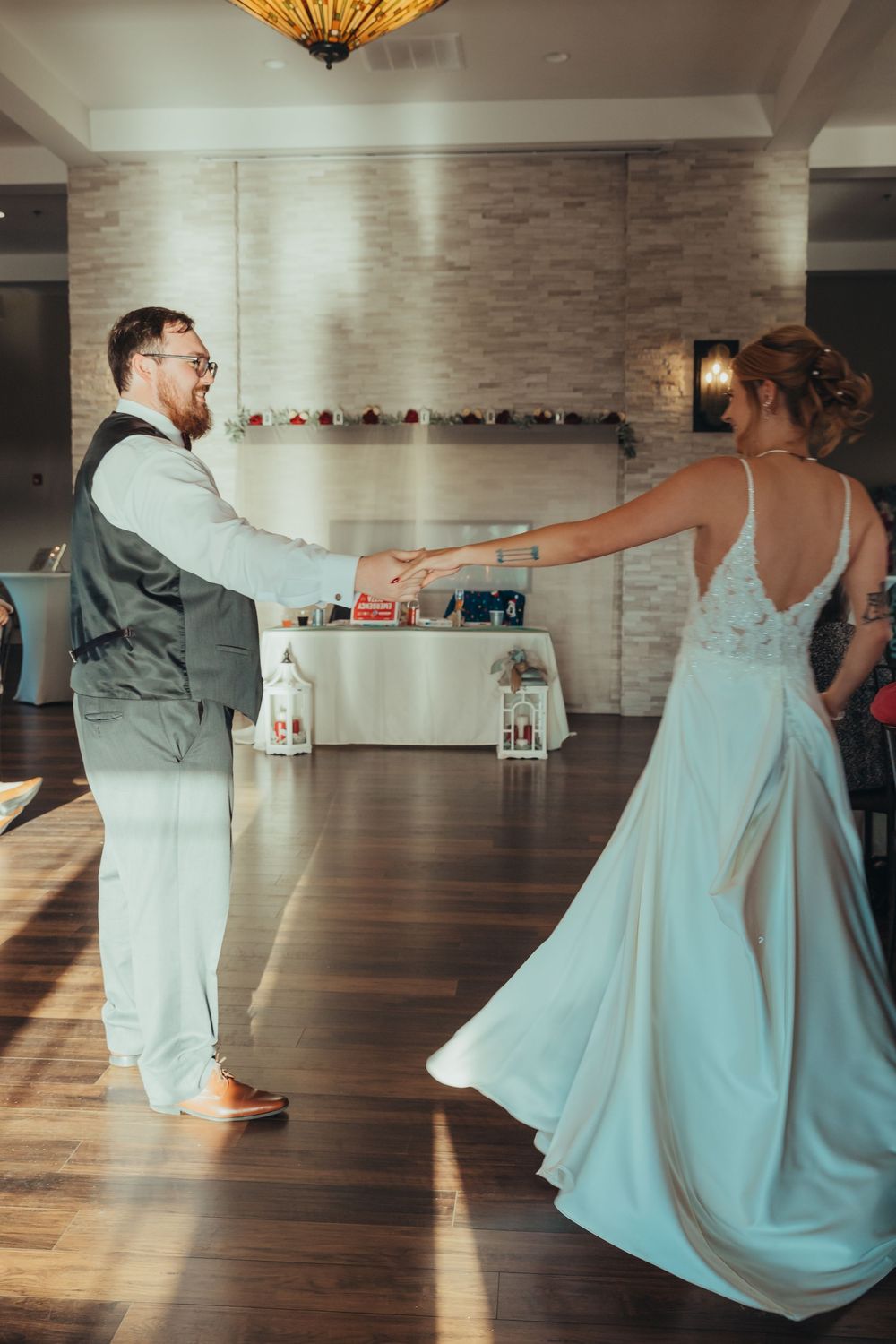 Two people dance at a wedding reception in formal attire on a wooden dance floor with a decorated wall backdrop.