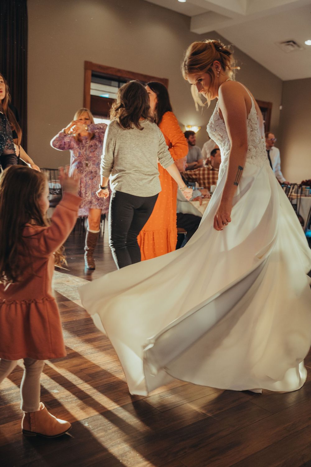 A person in a flowing white wedding dress twirls on a hardwood dance floor during a celebration.