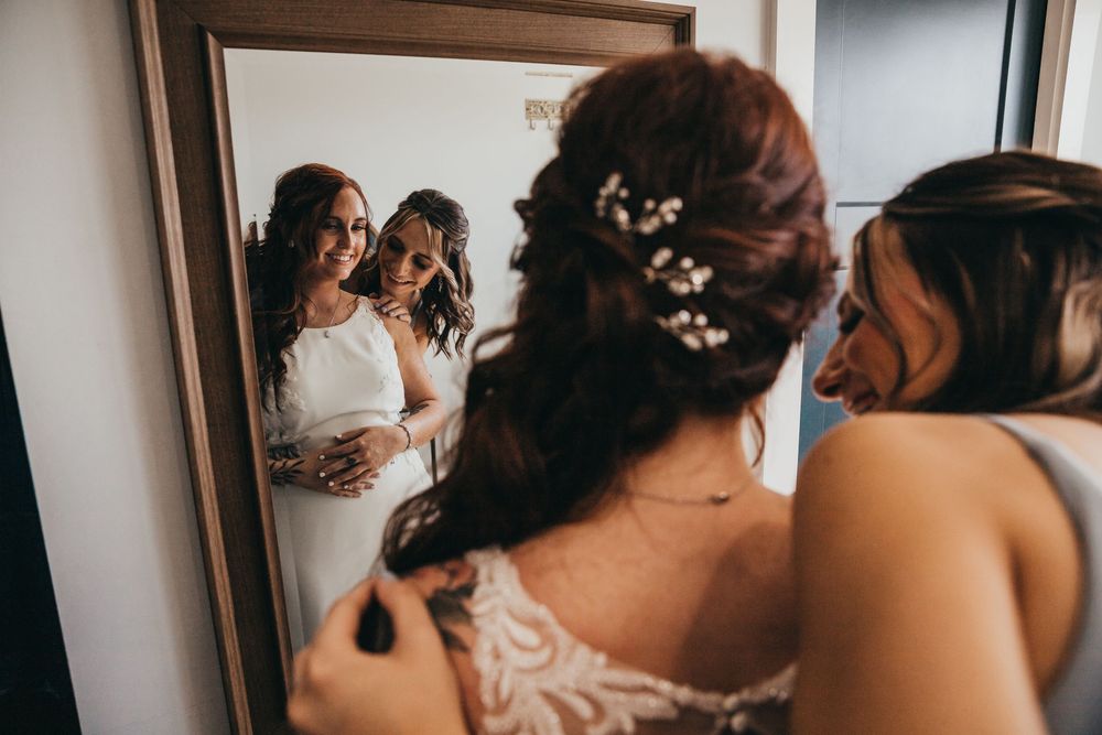 Reflection in mirror shows someone in a white lace wedding dress being helped with final preparations.