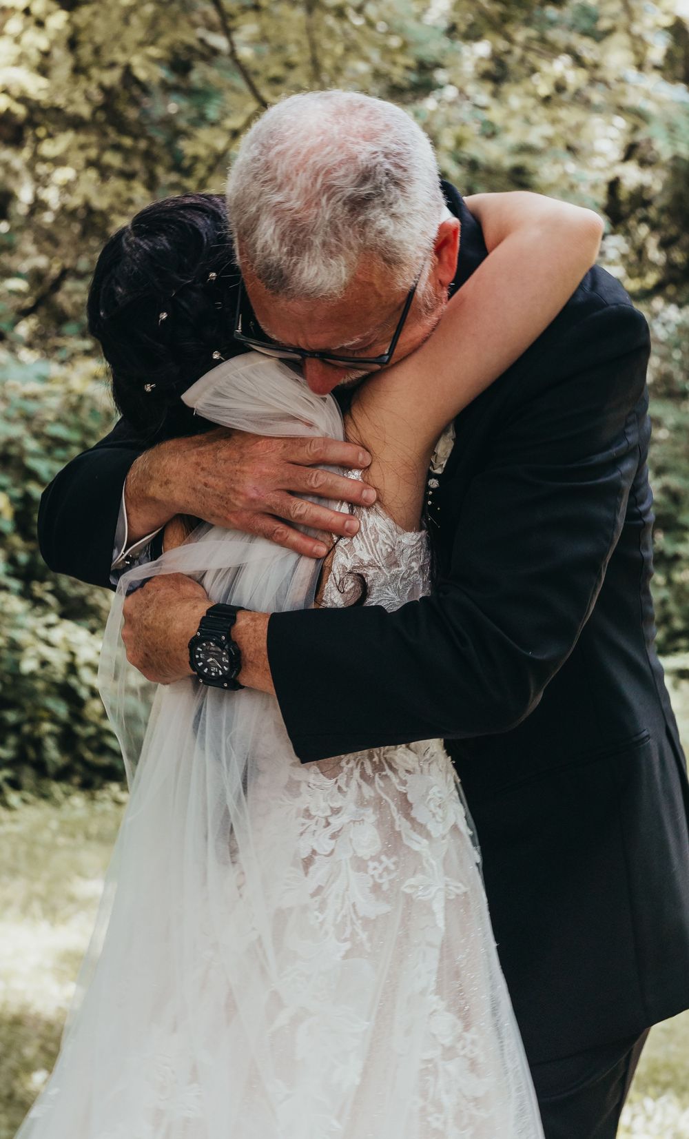 An emotional embrace between a man in a black suit and someone wearing a white lace wedding dress in an outdoor setting.