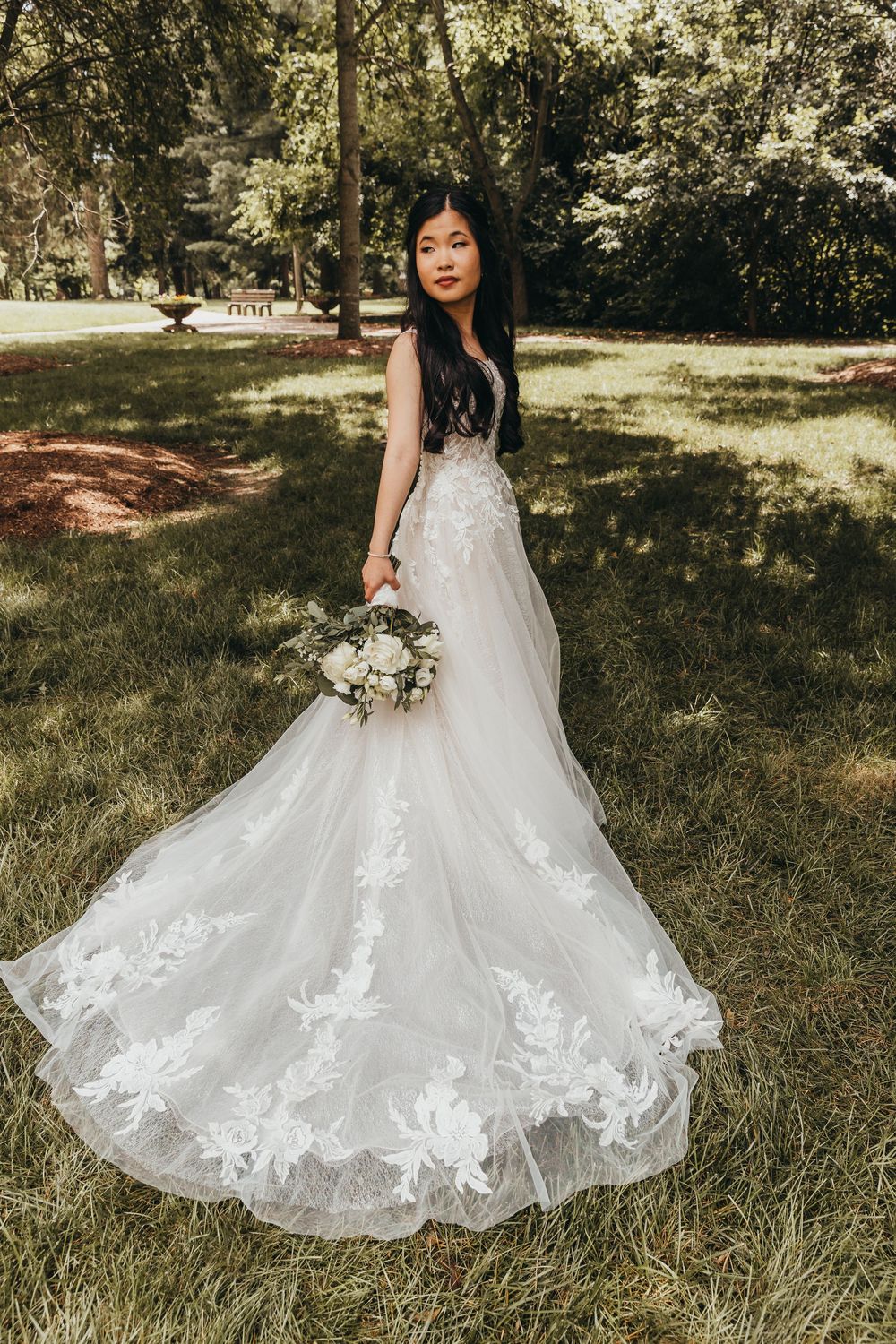 A bride in a stunning white lace wedding gown with a long train holds a bouquet in an outdoor garden setting.