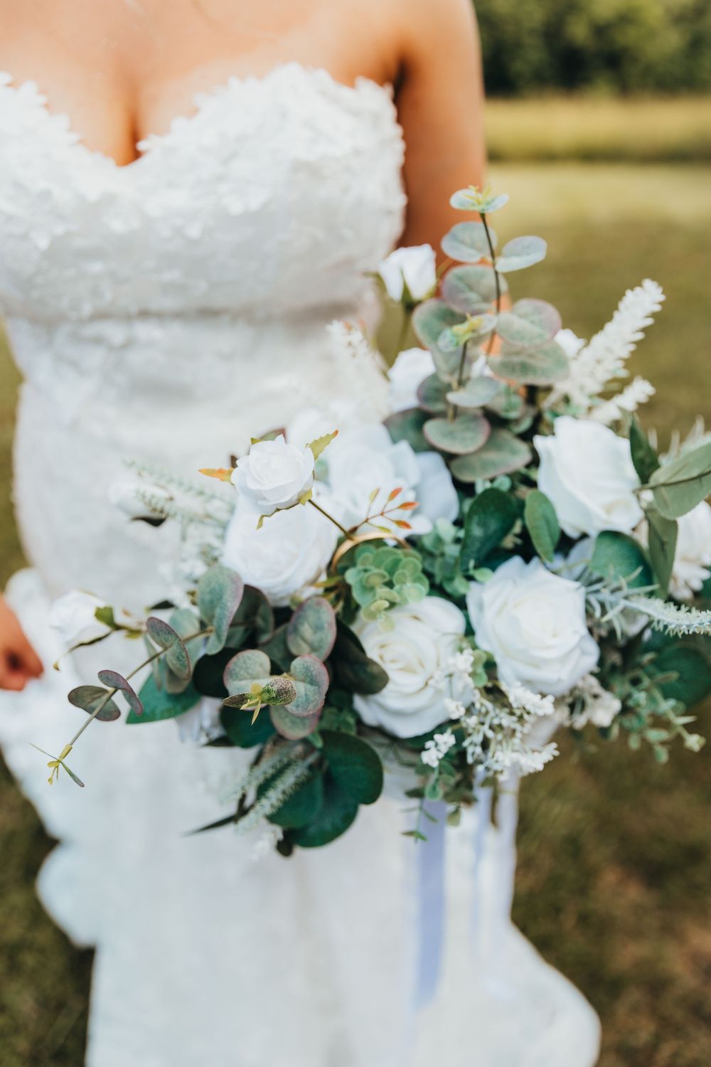 A wedding bouquet of white roses and eucalyptus leaves held against a white wedding dress in an outdoor setting.
